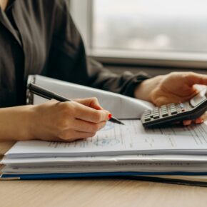 A close up of a female SR&ED tax incentive consultant holding a pen in one hand and a calculator. In front of her is a stack of papers on top of a desk. Image retrieved from Mikhail Nilov: https://www.pexels.com/photo/person-in-long-sleeve-shirt-holding-a-calculator-8297031/ for use by SR&ED Tax Consultant The InGenuity Group SR&ED Education and Resources